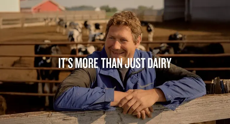 A smiling farmer leans on a fence with cows in the background and text saying "It's more than just dairy."