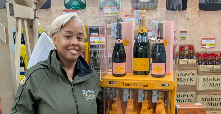 Une personne souriant à côté d'un présentoir de bouteilles de champagne Veuve Clicquot dans un magasin.