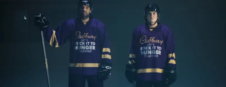 Two hockey players wearing purple jerseys with "Cadbury, Let's Stick It To Hunger Together" printed on them.