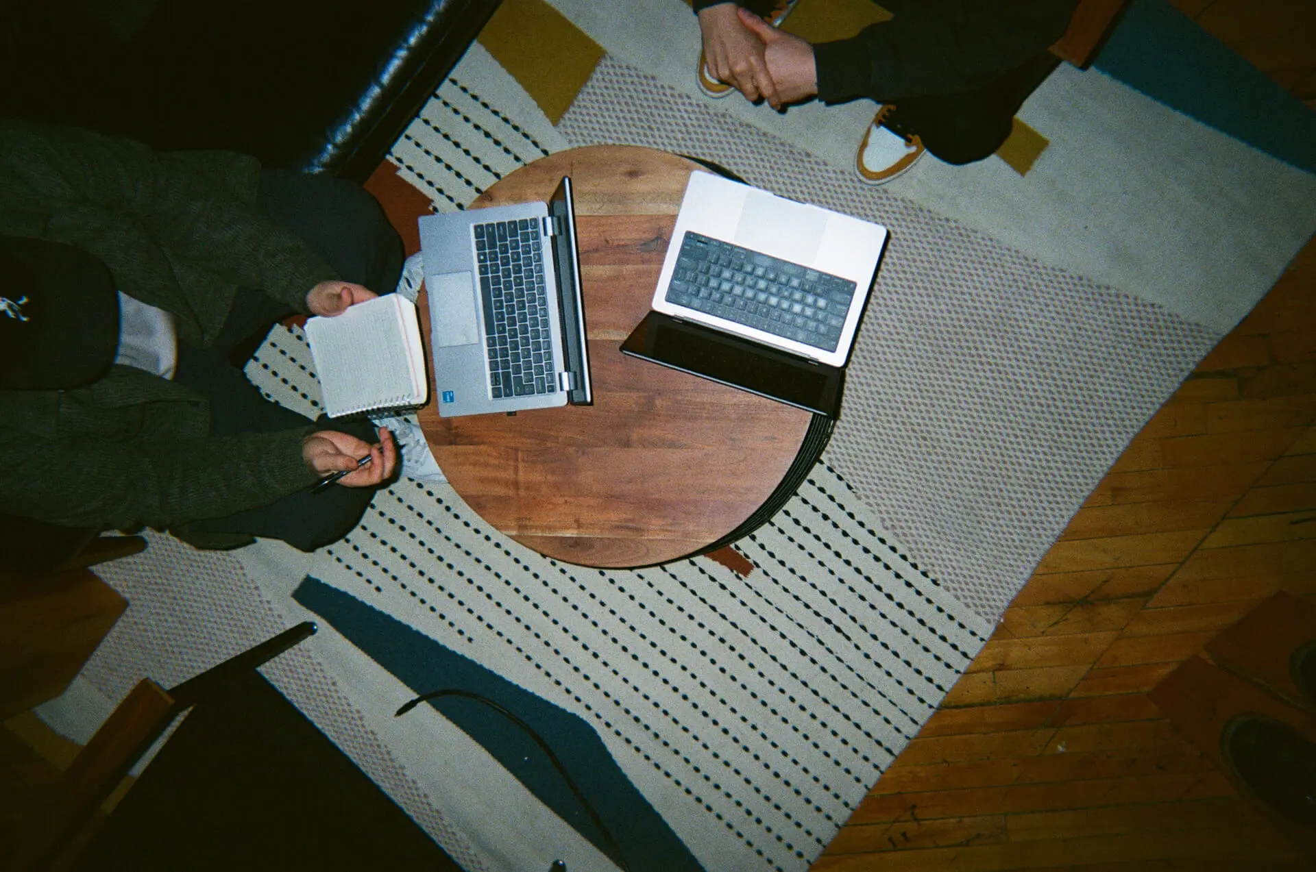 Vue aérienne de deux personnes assises à une table ronde avec des ordinateurs portables et un carnet sur un tapis à motifs.