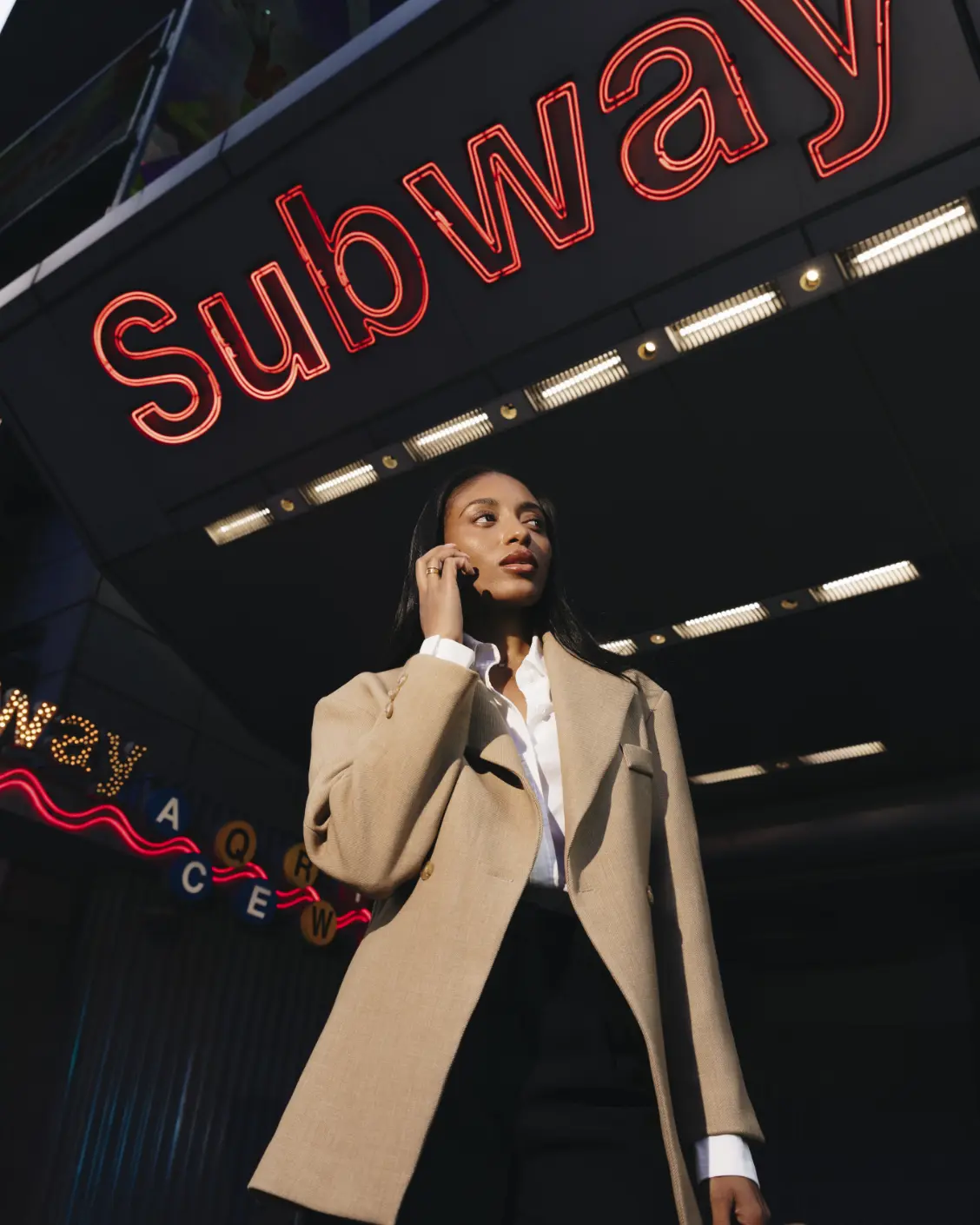 Une femme en manteau beige parle au téléphone sous un panneau de métro.