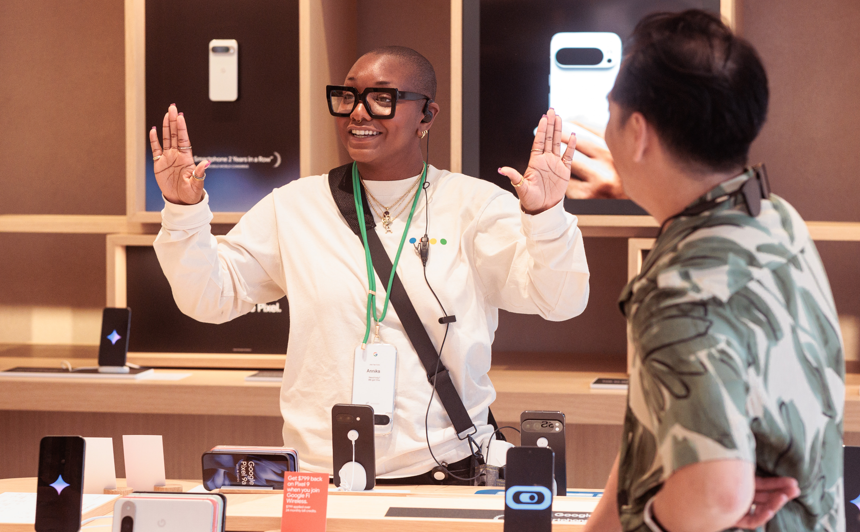 Une personne portant des lunettes et un badge parle avec enthousiasme à une autre personne dans un magasin de technologie.