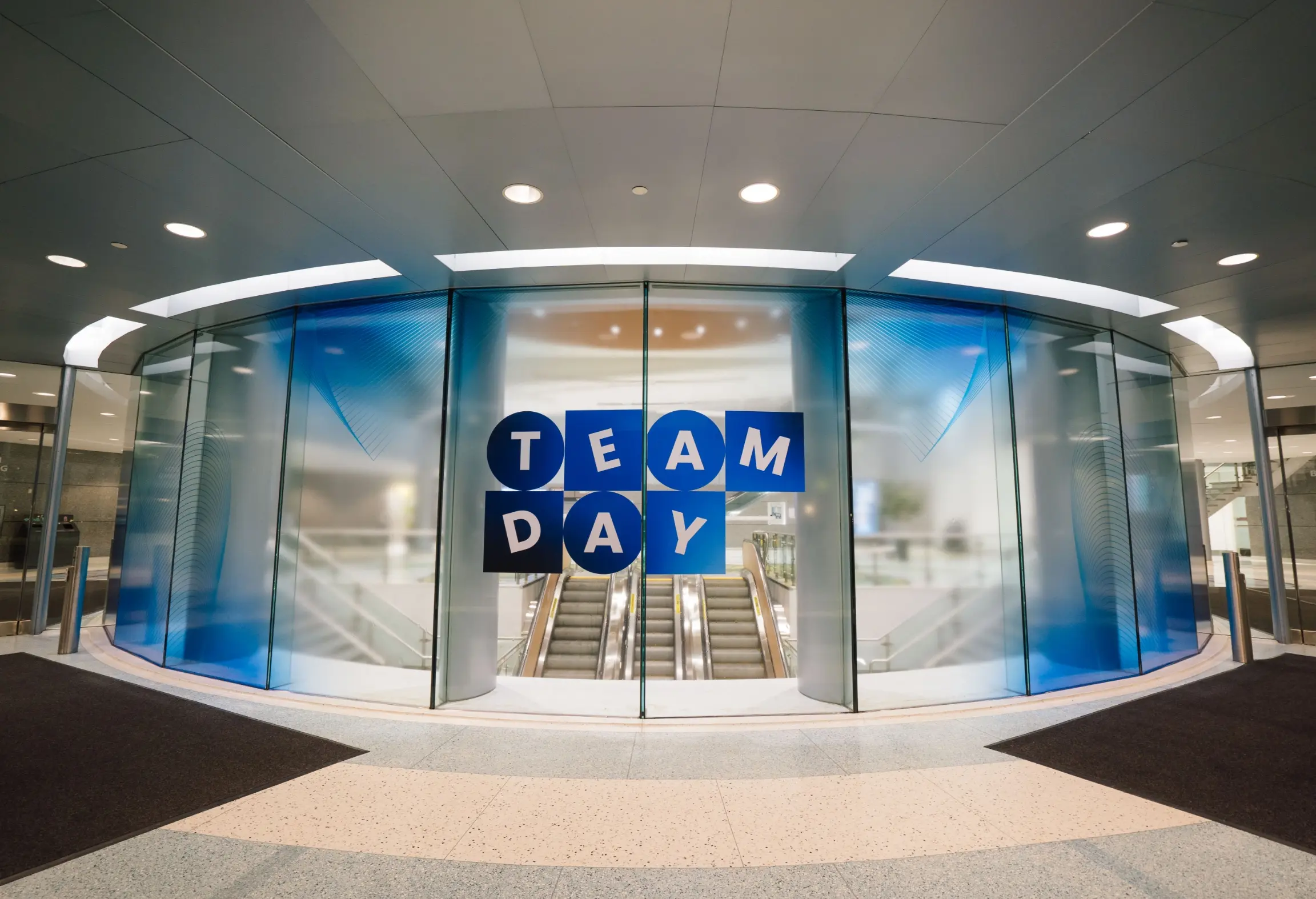 Des portes en verre avec "TEAM DAY" écrit en lettres bleues et blanches, menant à un escalier mécanique.