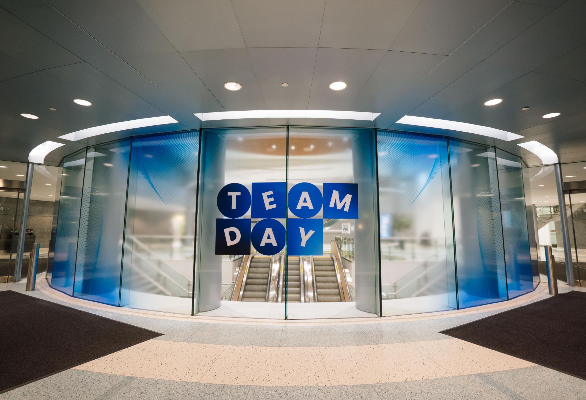 Des portes en verre avec "TEAM DAY" écrit en lettres bleues et blanches, menant à un escalier mécanique.