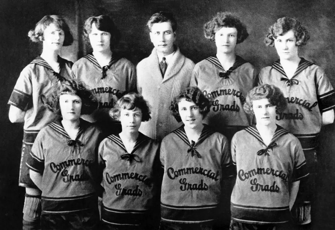 Une photo vintage en noir et blanc d'une équipe de basketball féminine et d'un entraîneur, avec « Commercial Grads » sur leurs uniformes.