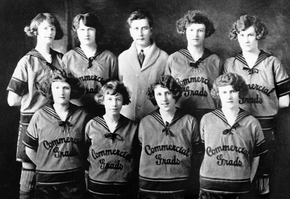 Une photo vintage en noir et blanc d'une équipe de basketball féminine et d'un entraîneur, avec « Commercial Grads » sur leurs uniformes.