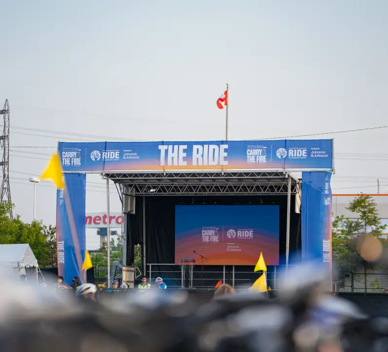 Outdoor stage with a large screen and "The Ride" banner, surrounded by a few people and yellow flags.