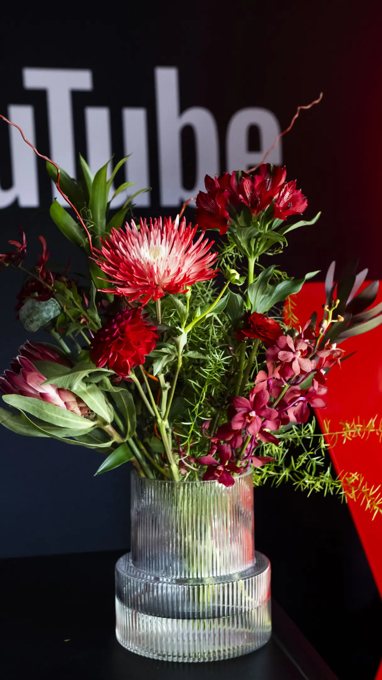 Un bouquet vibrant de fleurs rouges et roses dans un vase transparent nervuré, avec un logo partiellement visible en arrière-plan.