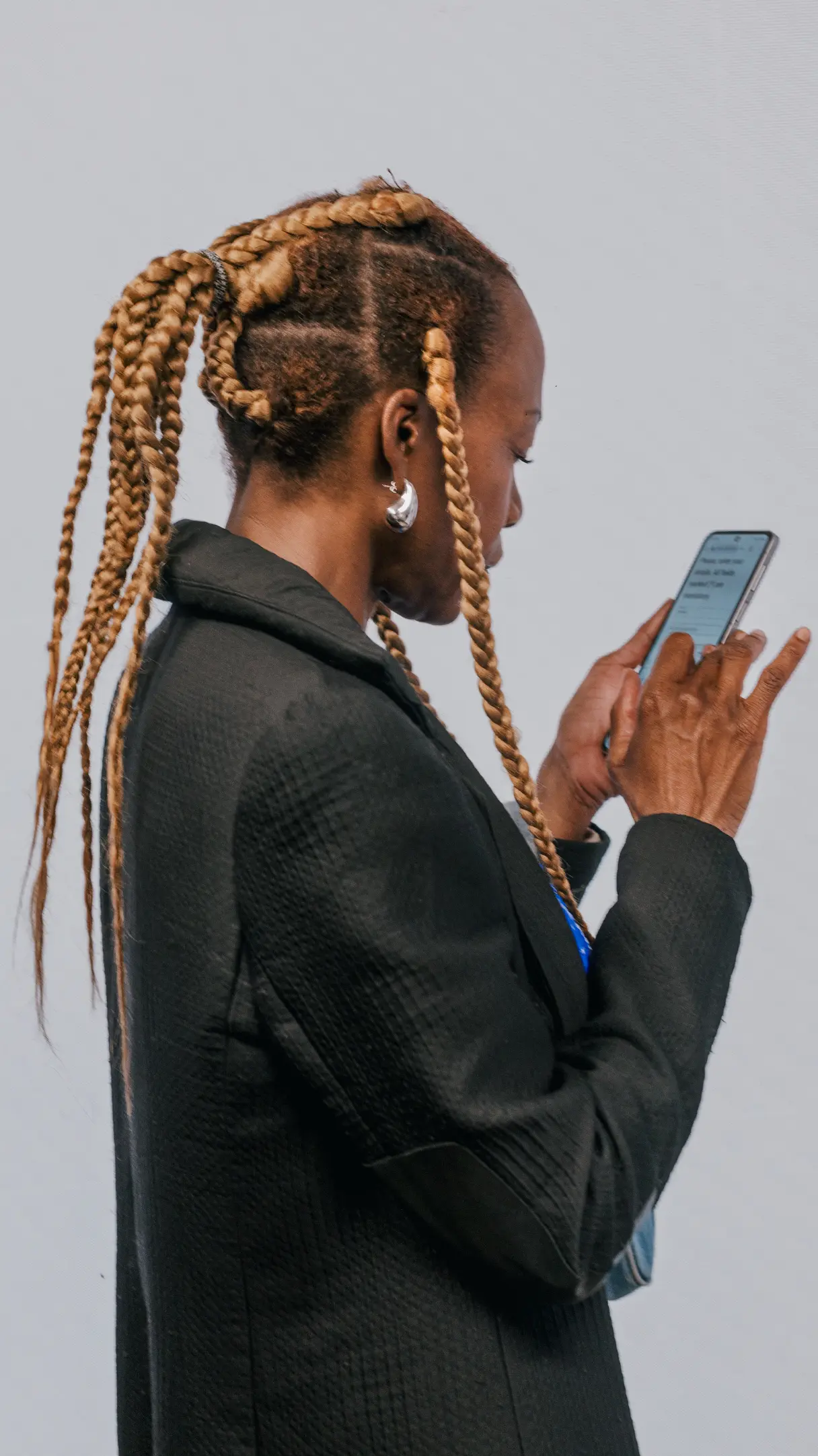Une personne aux cheveux tressés porte une veste noire et regarde un téléphone.
