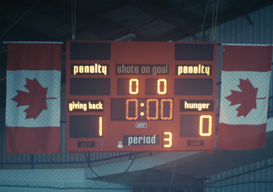 A hockey scoreboard shows "Giving Back" leading "Hunger" 1-0 with 0:00 remaining in the third period, flanked by Canadian flags.