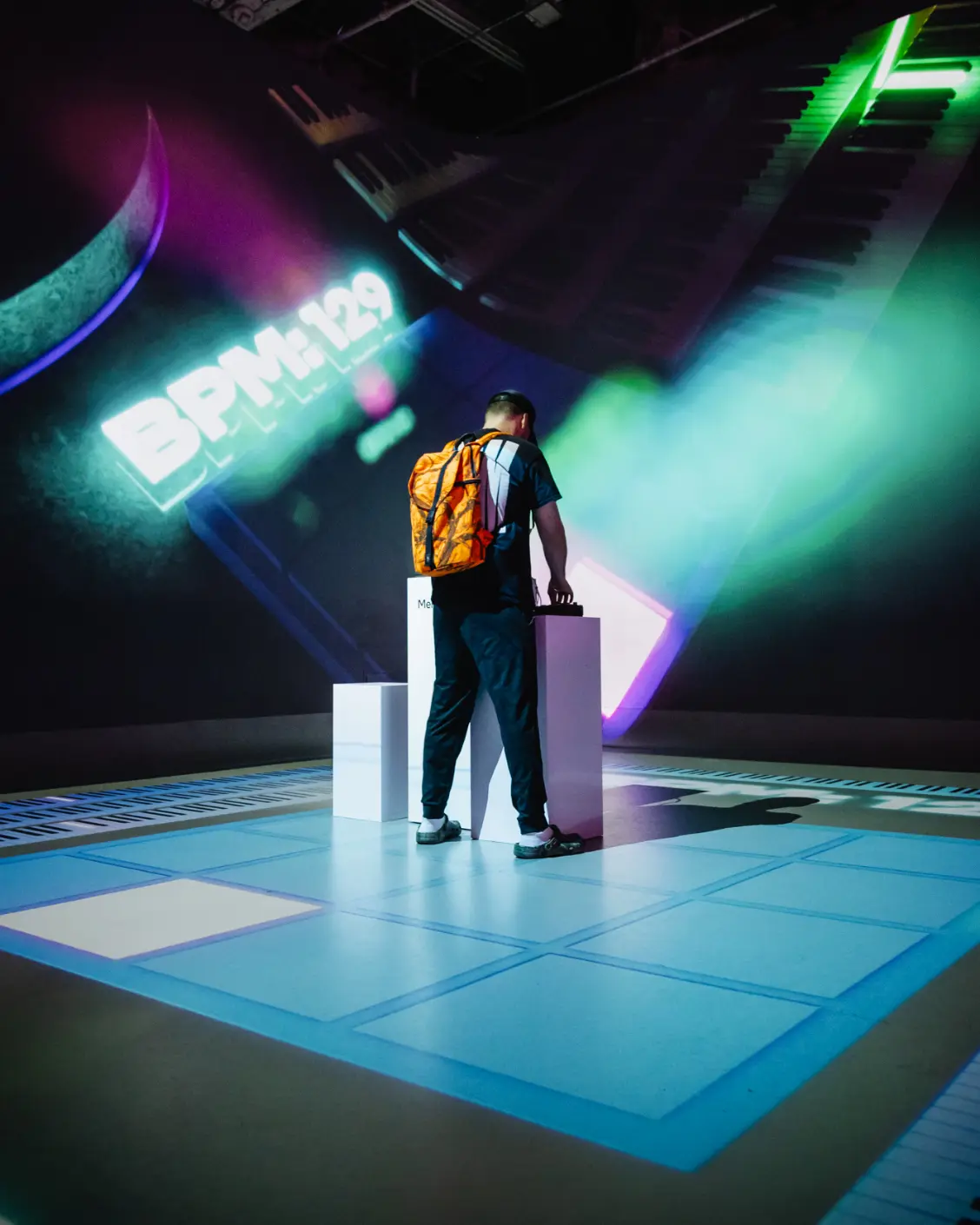 Une personne avec un sac à dos orange interagit avec une exposition sous des lumières colorées, avec « BPM: 129 » affiché en arrière-plan.