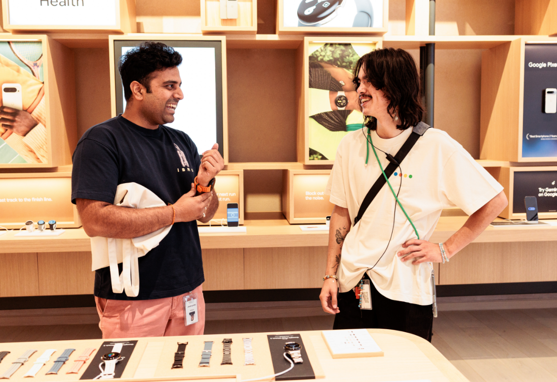 Deux personnes souriant et parlant dans un magasin de technologie près d'un présentoir de montres intelligentes et d'accessoires.