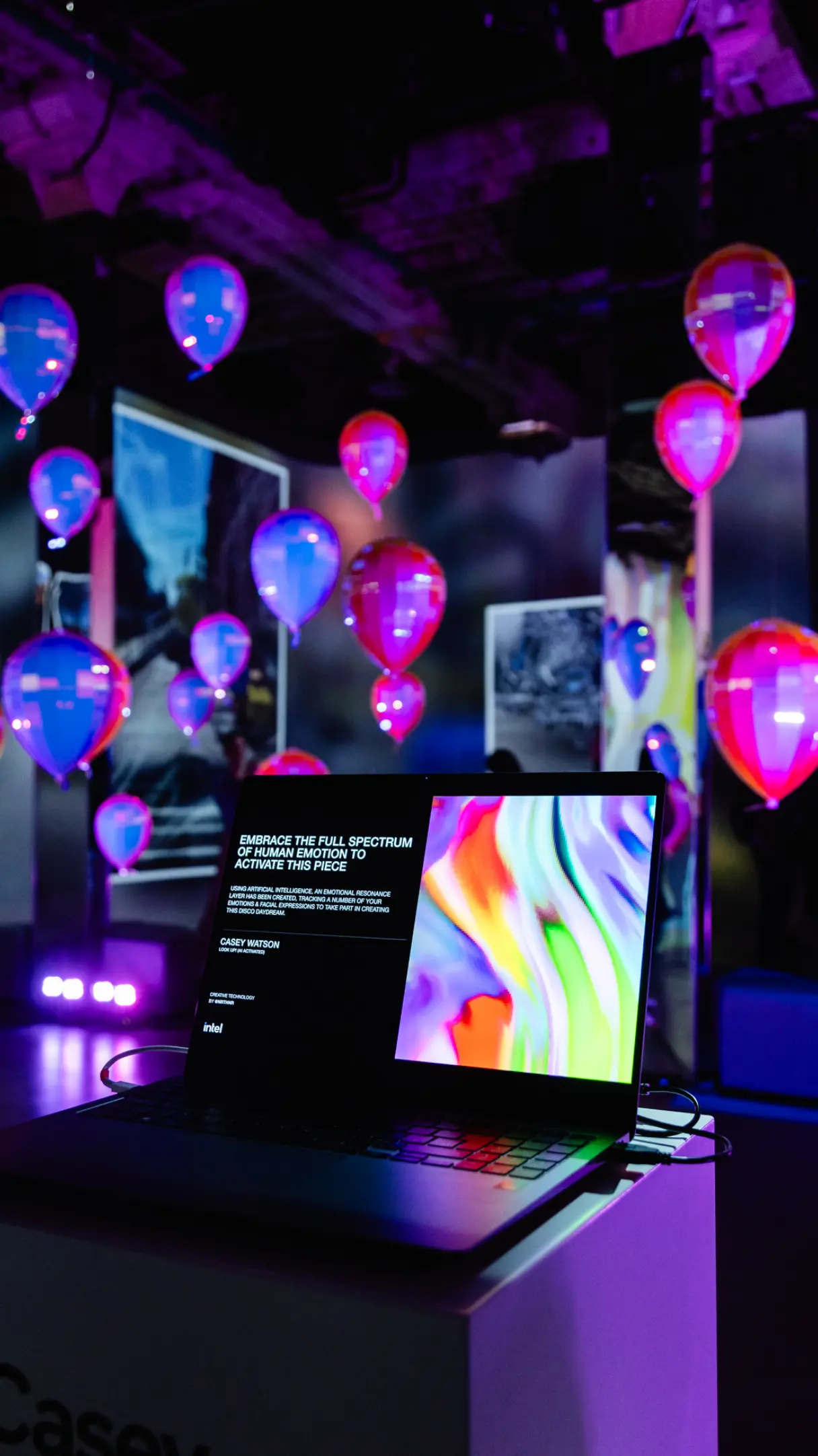 Un ordinateur portable affichant de l'art numérique coloré est installé dans une pièce avec des ballons lumineux et des lumières vibrantes.