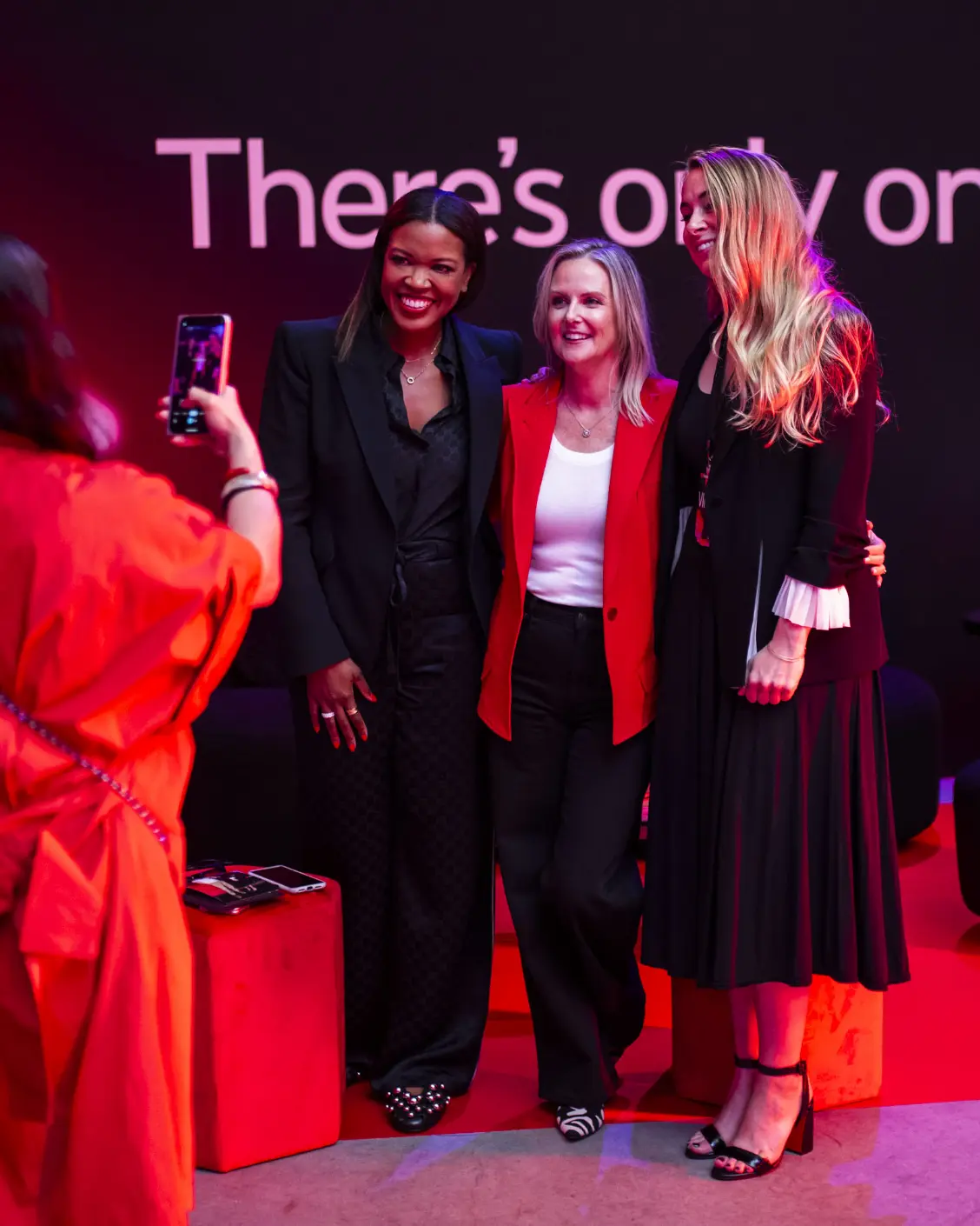Trois femmes posent pour une photo ensemble lors d'un événement avec un panneau « There's only on » en arrière-plan.