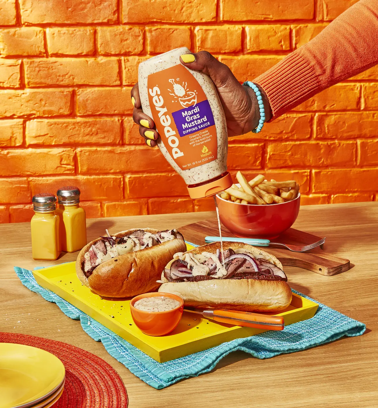 Une personne verse de la sauce Mardi Gras Mustard sur des sandwichs sur une table en bois, à côté de frites et de condiments.