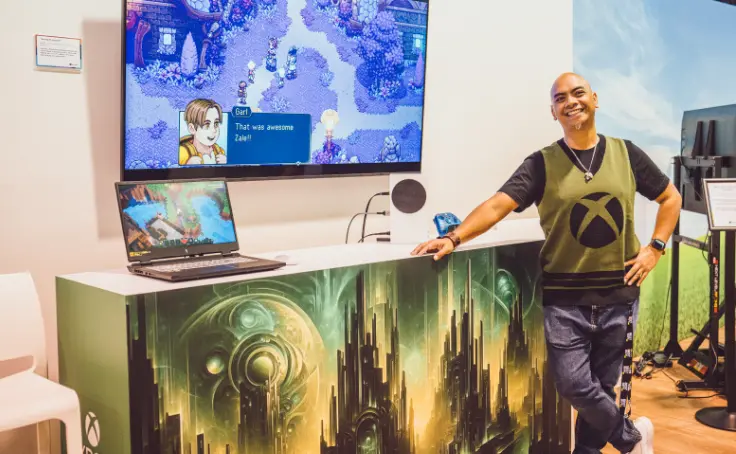 A person smiling and standing next to a gaming setup with a video game displayed on a monitor and laptop.