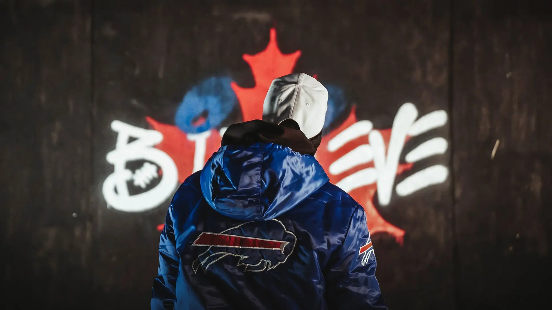 Une personne en veste bleue avec un emblème de buffle se tient devant un graffiti représentant une feuille d'érable.