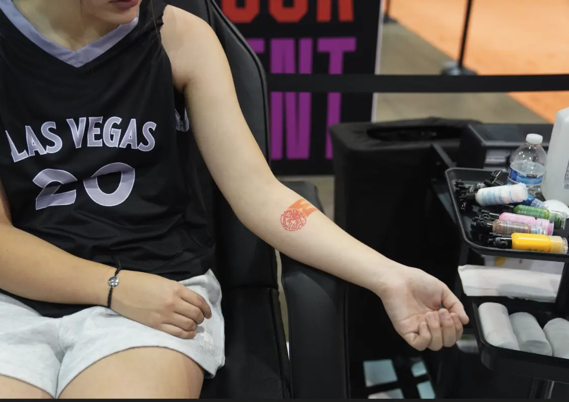 Une personne portant un maillot « Las Vegas » affiche un tatouage temporaire rouge sur son bras tout en étant assise à côté d'un plateau de fournitures de tatouage.