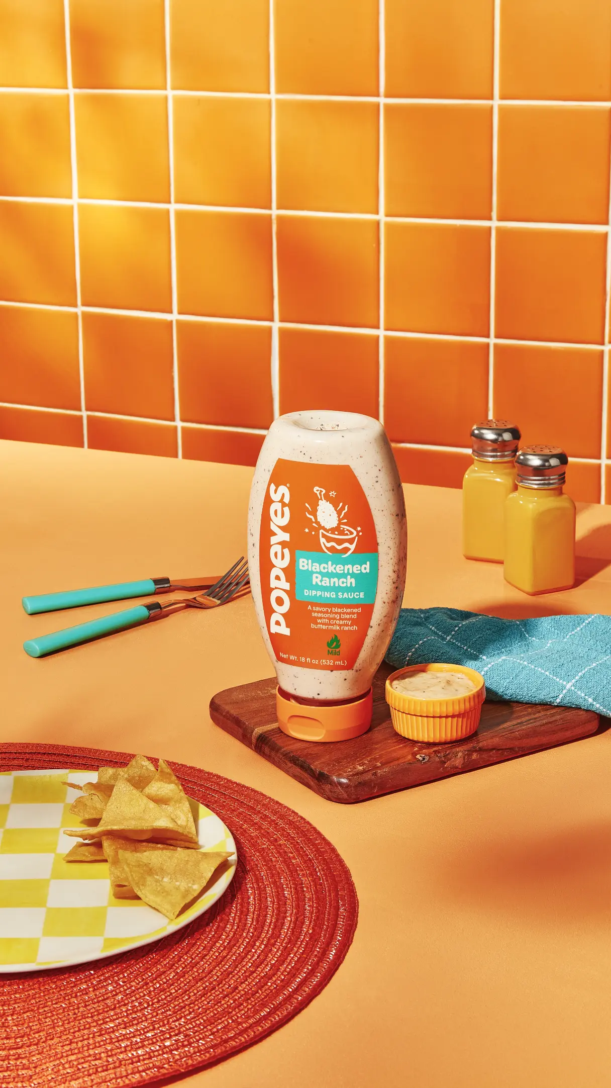 A bottle of Popeyes Blackened Ranch dipping sauce on a table with chips, utensils, and condiments against an orange tiled background.