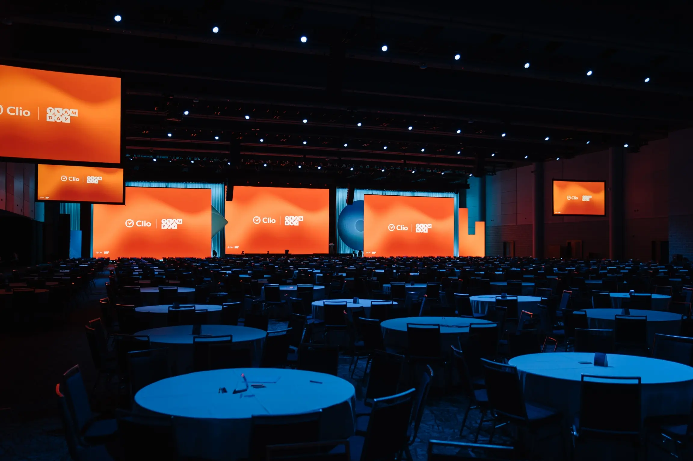 Une grande salle de conférence faiblement éclairée avec plusieurs tables rondes et des écrans orange vif affichant "Clio" et "Team Day."