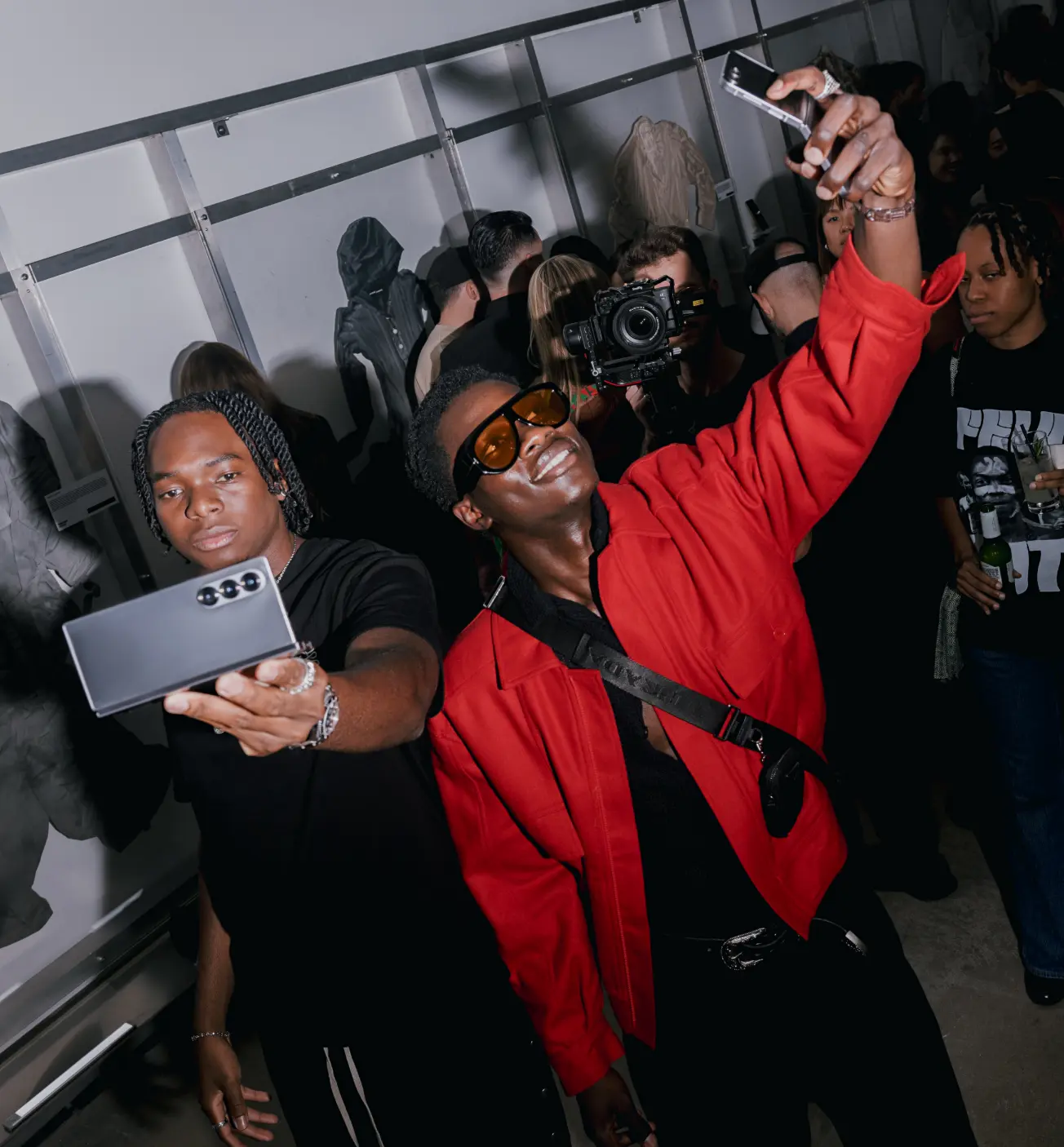 Deux personnes prennent des selfies lors d'un événement bondé, avec d'autres personnes en arrière-plan, dont quelqu'un tenant une caméra.