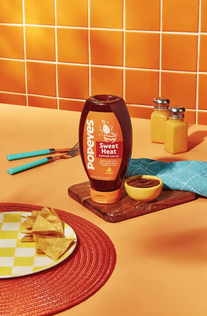 Une bouteille de sauce trempette Popeyes Sweet Heat est placée sur une planche en bois à côté d'un petit bol de sauce, avec des chips de tortilla sur une assiette à carreaux et un fond de carreaux orange.