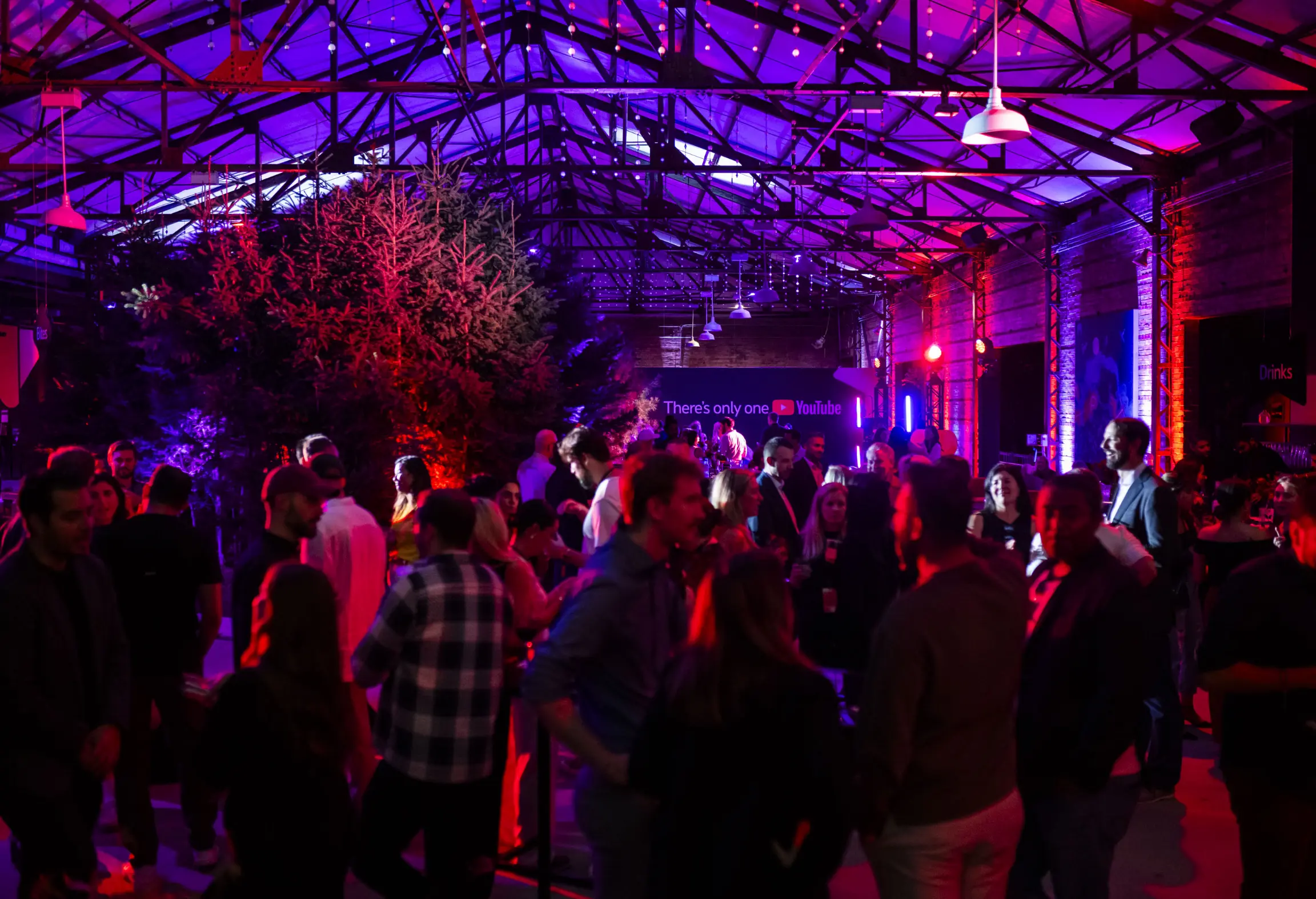 A lively indoor party with colorful lighting, featuring a crowd of people socializing under a high ceiling.