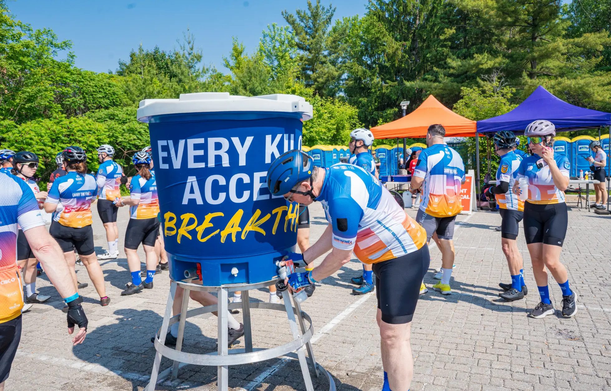 Cyclists wearing matching jerseys gather around a large blue water cooler during a bike event.