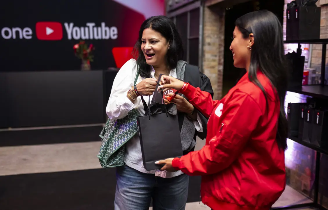 Two women smiling and exchanging a black gift bag, with a YouTube logo in the background.