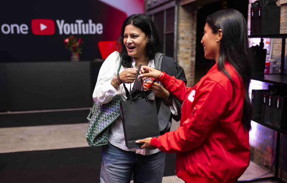 Deux femmes souriant et échangeant un sac cadeau noir, avec un logo YouTube en arrière-plan.