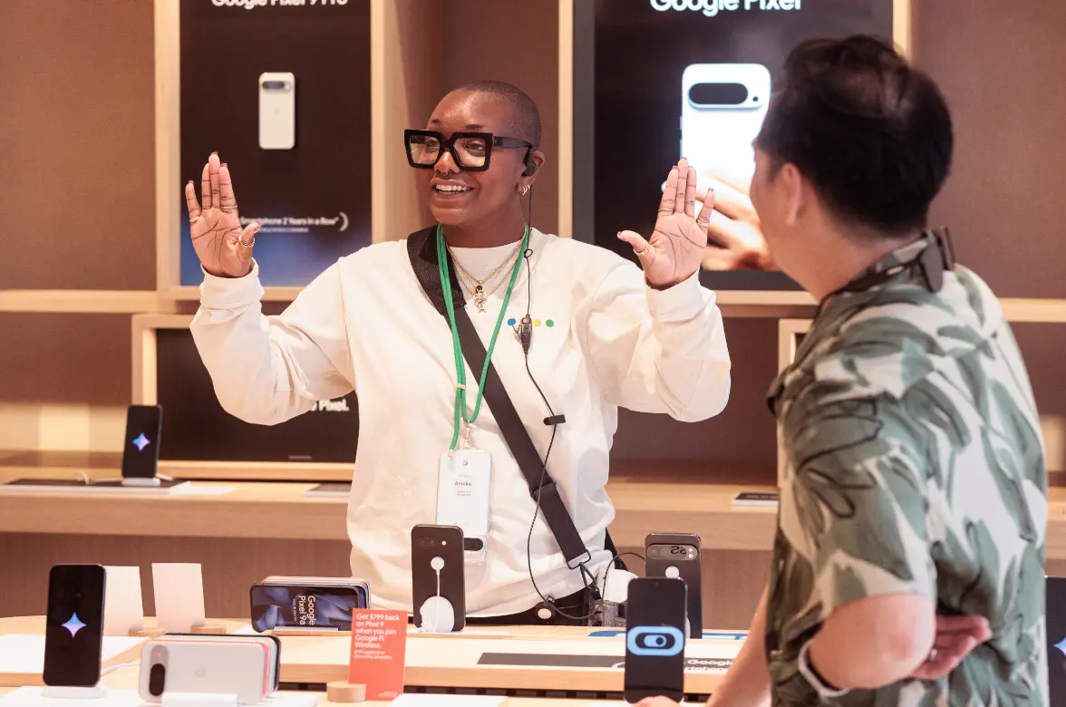 Une personne portant une chemise blanche et des lunettes parle avec enthousiasme à une autre personne dans un magasin exposant des appareils Google Pixel.
