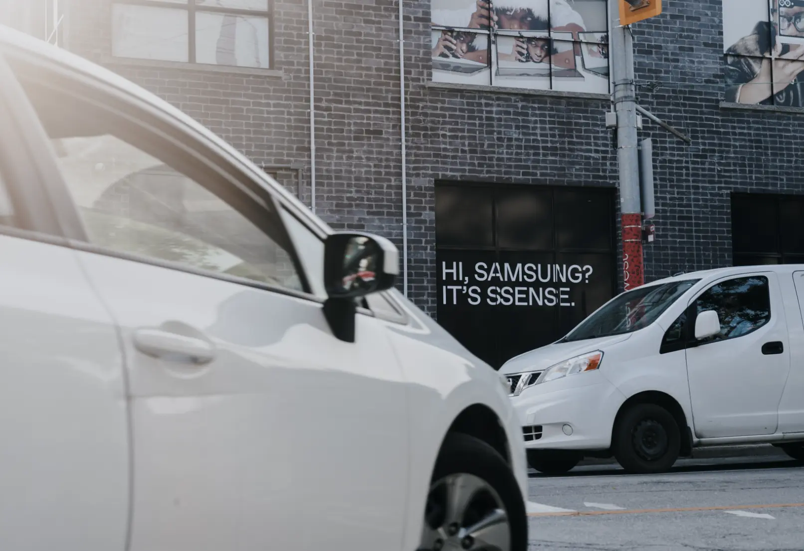 Des voitures garées dans une rue devant un bâtiment en briques avec un message disant « Salut, Samsung ? C'est SSENSE. »