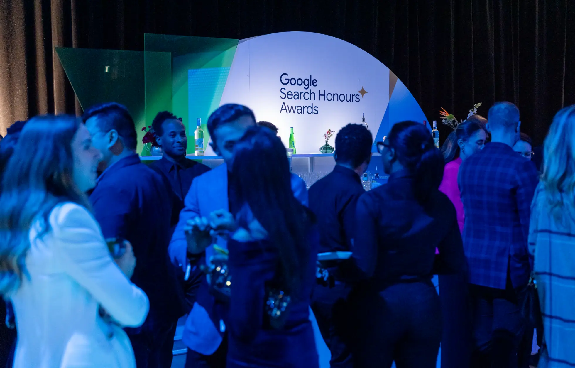 A group of people socializing at an event with a sign reading "Google Search Honours Awards" in the background.