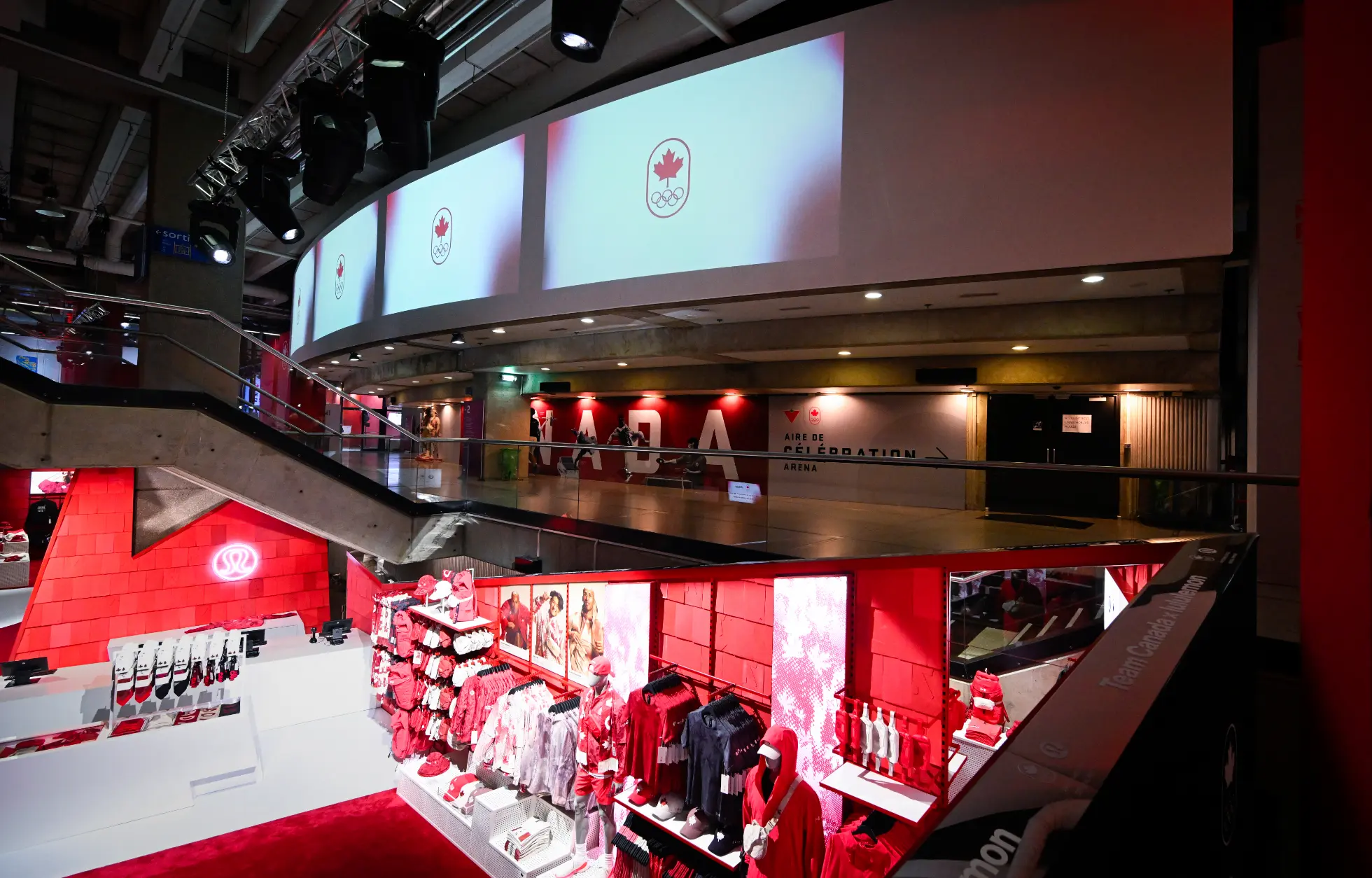 Un magasin de détail avec des présentoirs de marchandises rouges sous de grands écrans montrant un symbole olympique canadien.