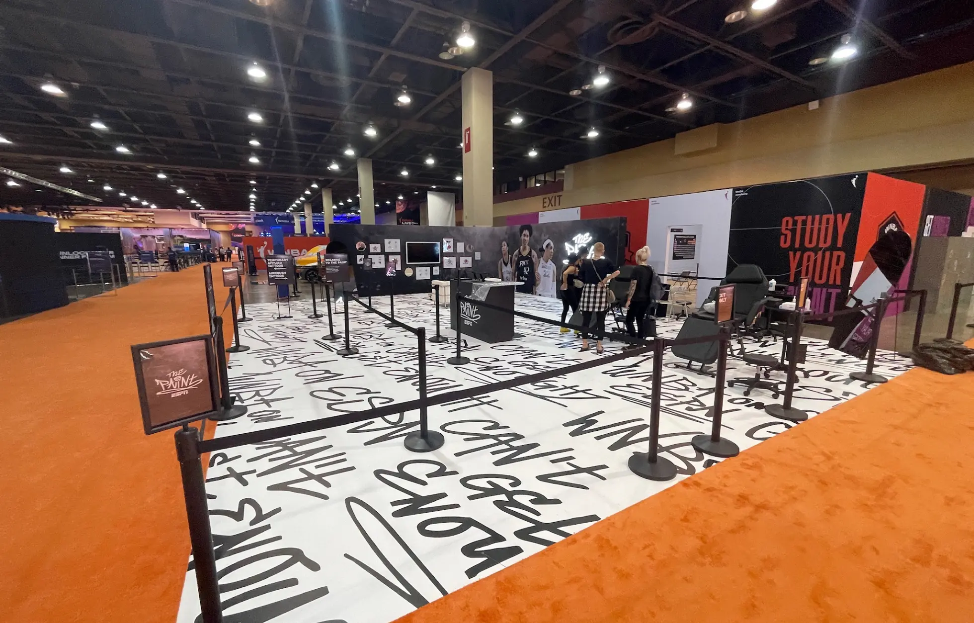Zone spacieuse de centre de congrès avec des installations artistiques, une impression au sol en noir et blanc, et des personnes interagissant à un stand d'exposition.