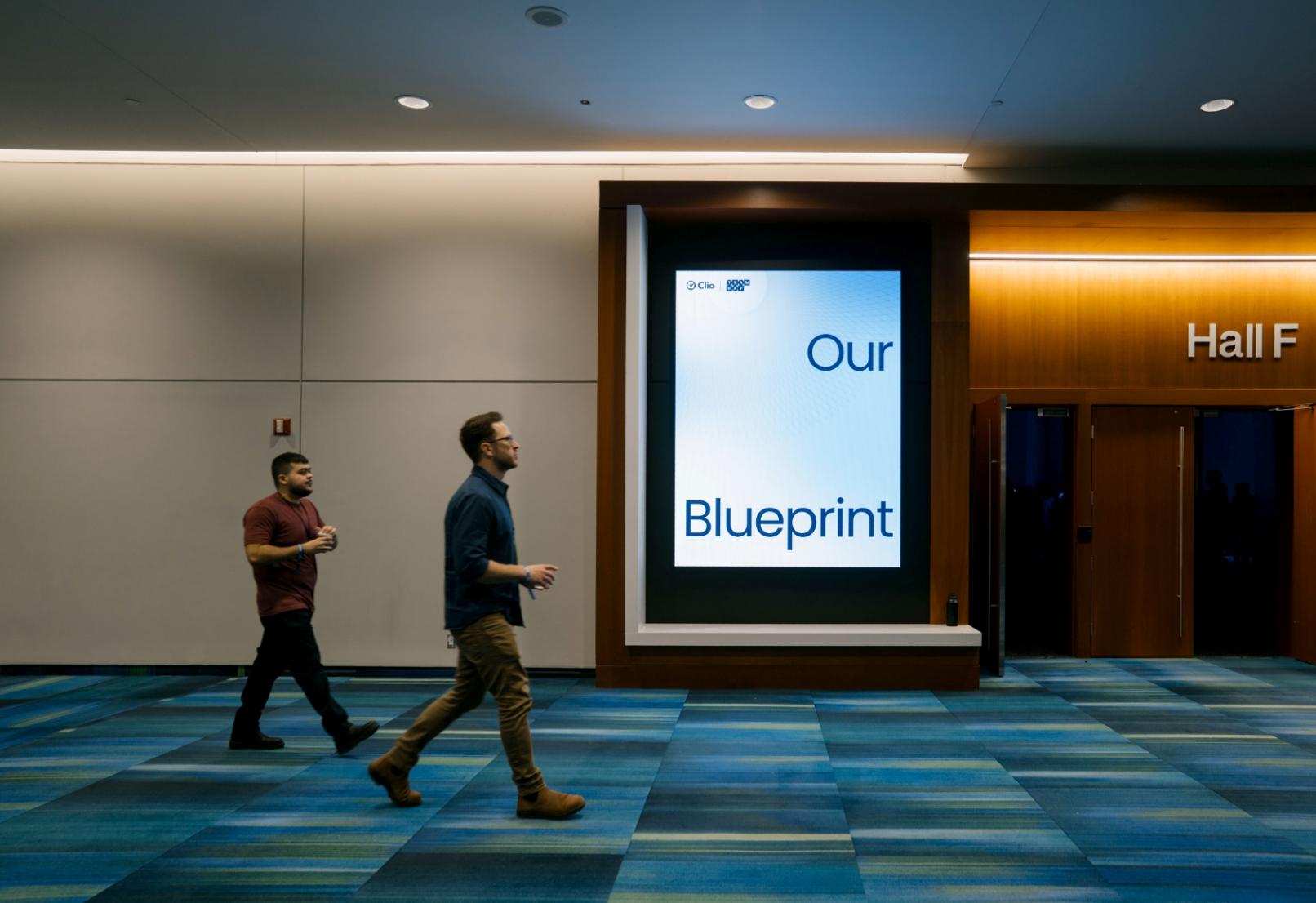 Deux personnes marchant dans un couloir avec un panneau numérique indiquant "Our Blueprint" et des portes vers la salle F.