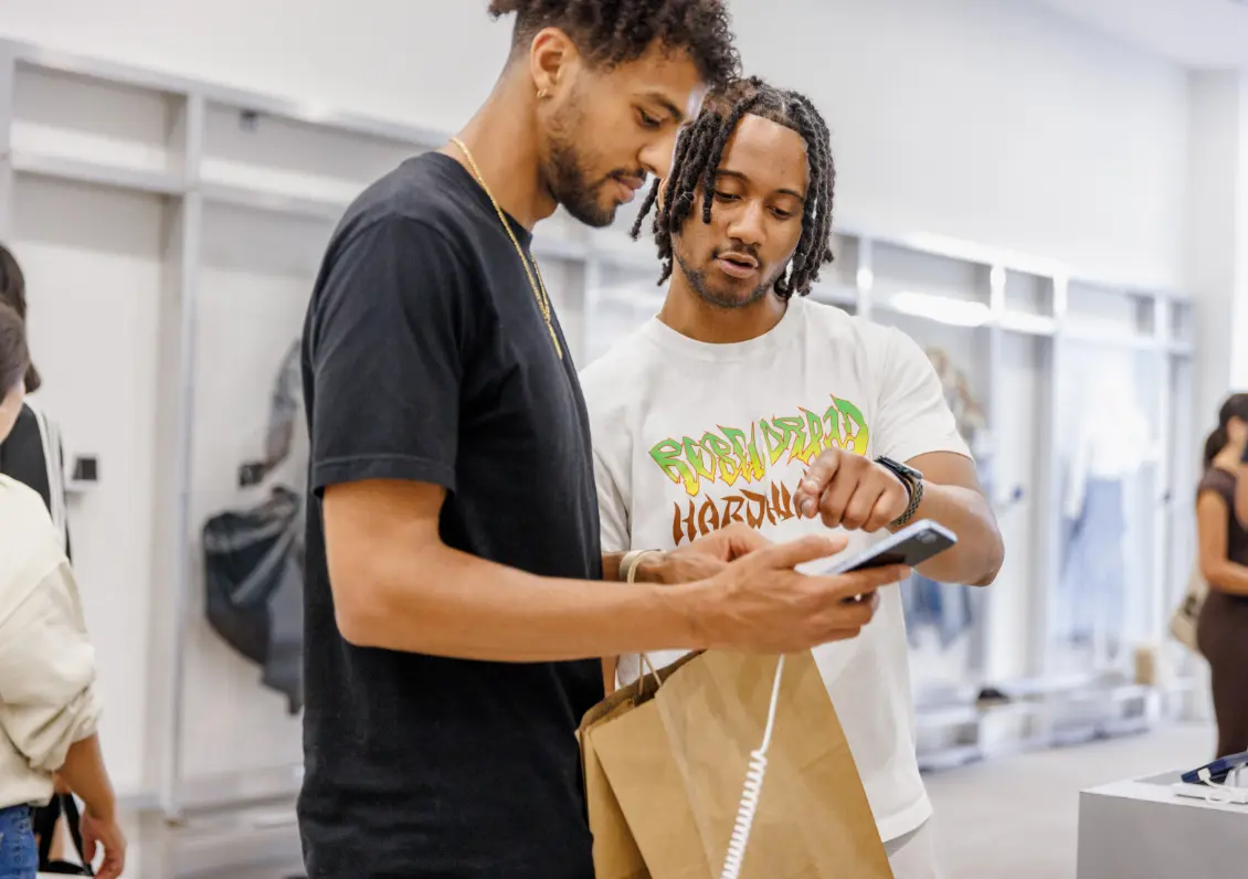 Deux hommes regardent ensemble un téléphone dans un magasin, l'un tenant un sac de courses.