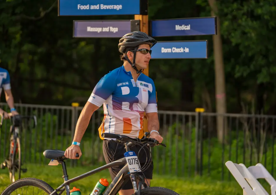 A cyclist wearing a helmet stands with a bike near event signs outdoors.
