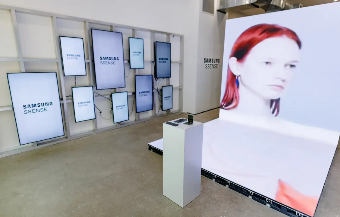 Un espace d'exposition présente plusieurs écrans Samsung avec des logos et un grand écran montrant une femme aux cheveux roux.