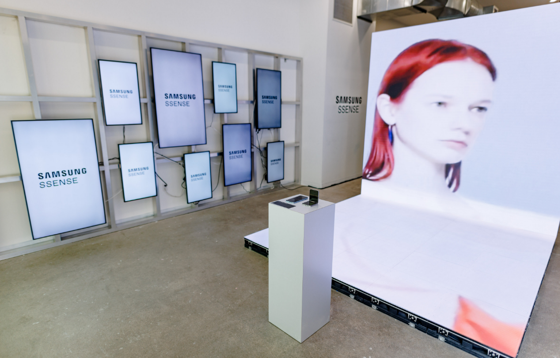 Un espace d'exposition présente plusieurs écrans Samsung avec des logos et un grand écran montrant une femme aux cheveux roux.