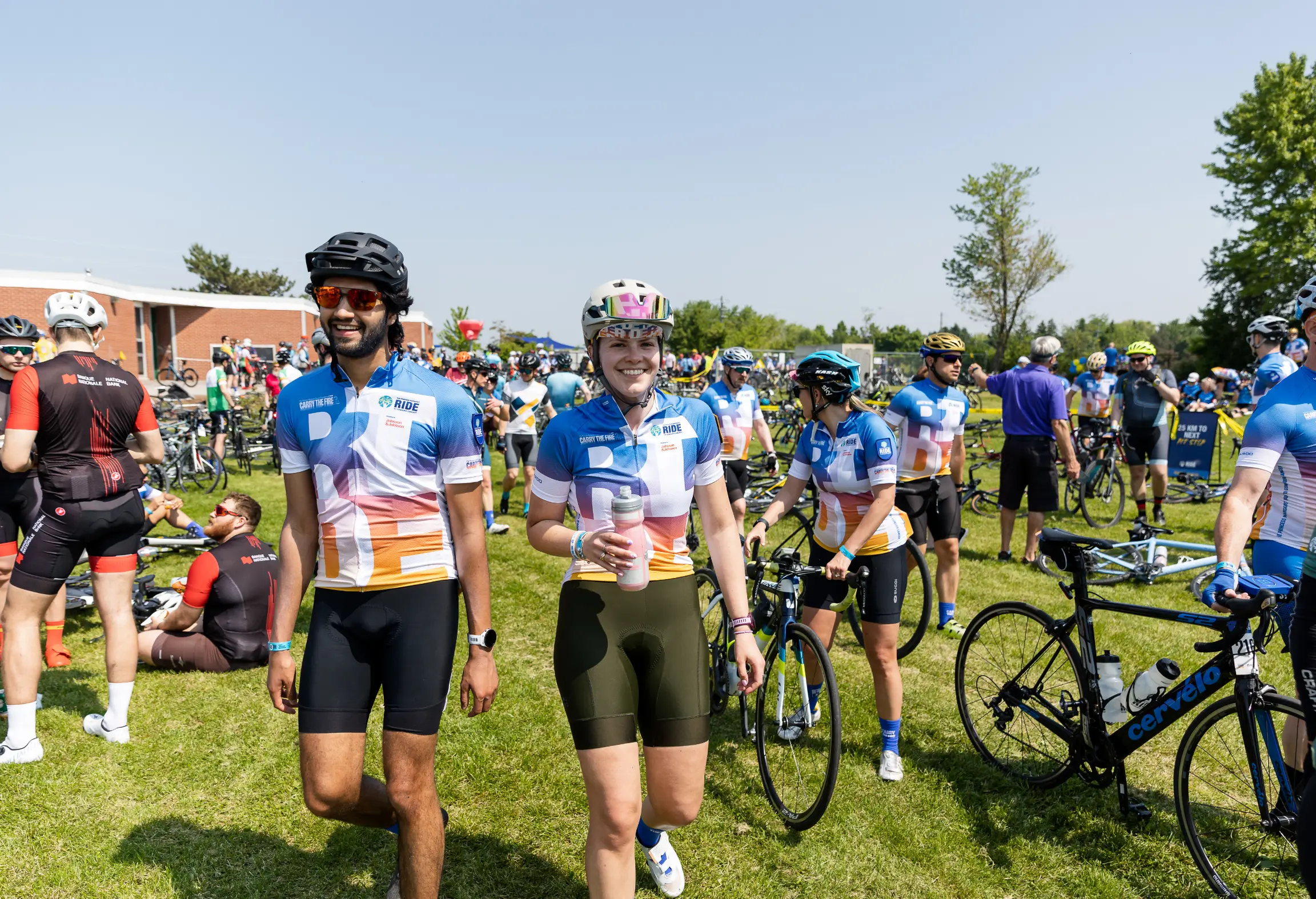 Cyclists in colorful jerseys gather on a grassy field, some holding bikes and others standing or sitting.