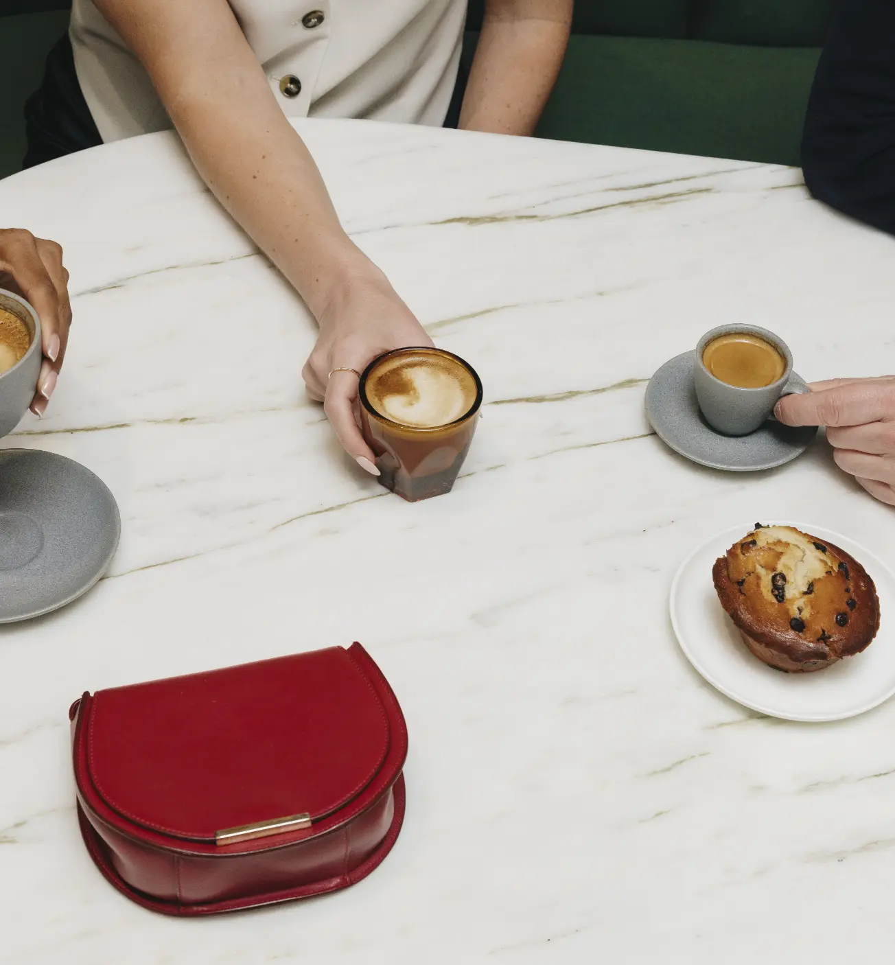 Trois personnes savourant du café et un muffin à une table en marbre avec un sac à main rouge posé à proximité.