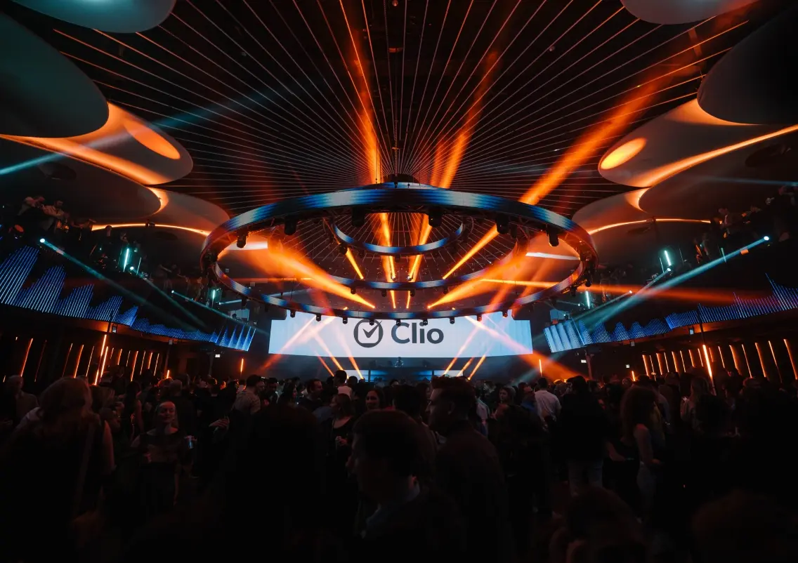 Une scène de boîte de nuit animée avec une foule, un éclairage coloré et un grand écran affichant le nom "Clio."