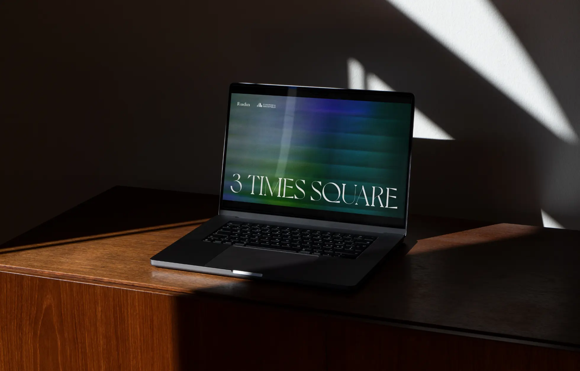 Un ordinateur portable affichant « 3 Times Square » sur son écran est posé sur une surface en bois avec un éclairage d'ombre dramatique.