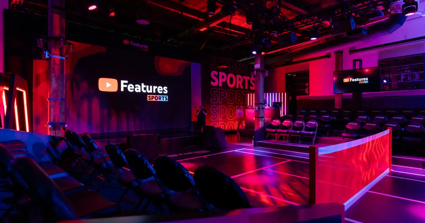 Un espace événementiel faiblement éclairé avec une scène affichant « Features Sports » sur un grand écran, entouré de rangées de sièges.