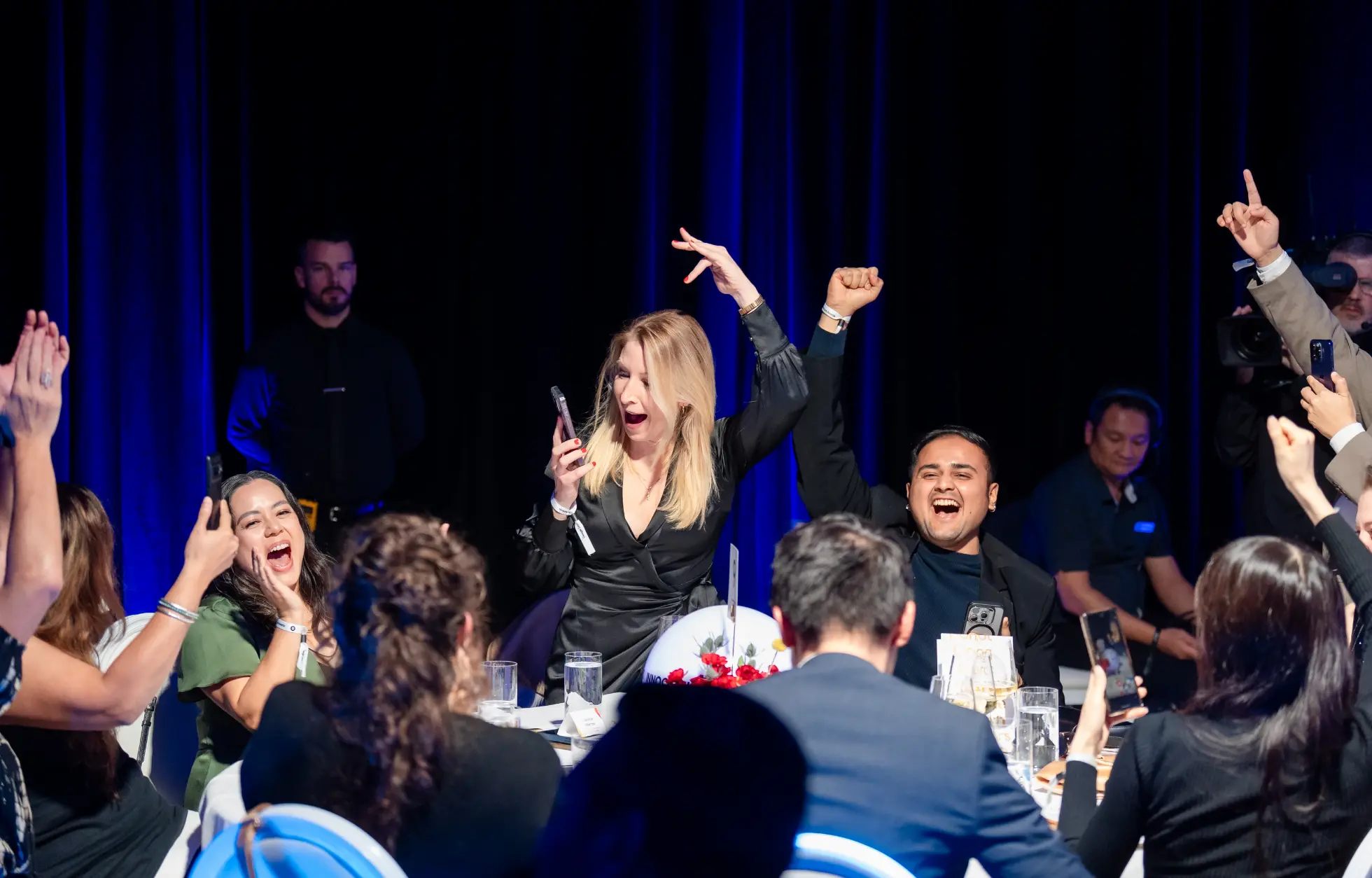 People at a table cheer and celebrate, some holding their phones up.