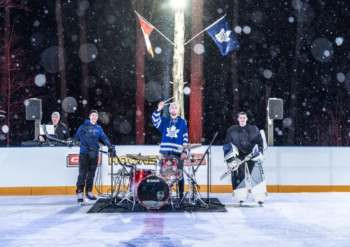 Trois personnes debout sur une patinoire de hockey extérieure dans la neige, l'une jouant de la batterie.