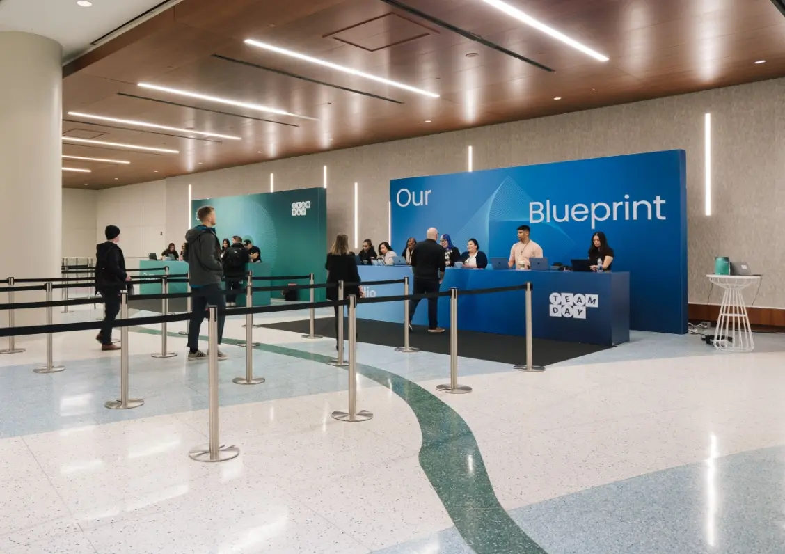 Des personnes sont rassemblées à un bureau d'inscription moderne avec une toile de fond bleue "Our Blueprint."