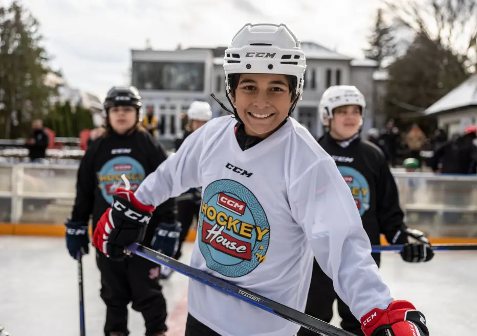 Un jeune joueur de hockey en maillot blanc sourit sur une patinoire extérieure avec des coéquipiers en arrière-plan.